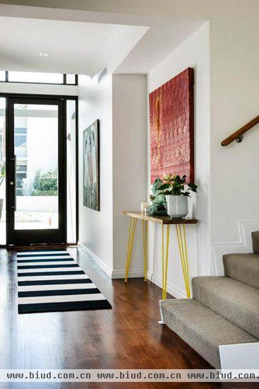 木框玻璃门设计从视觉上使室内外空间相连，而且方便自然光的透入。走在黑白条纹的地毯上就像踩着黑白钢琴键一样，让人心情愉悦，与桌子的黄色支架和楼梯级搭配在一起，使玄关处充满线条美。