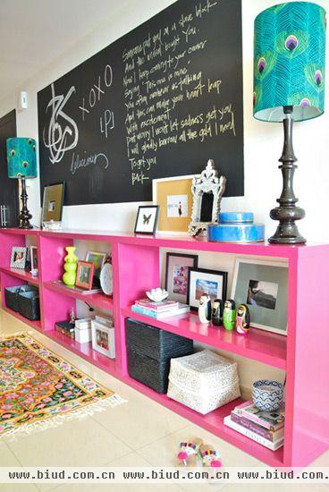 粉红色的储物架靠墙放置，不仅实现了物品收纳的功能，也美化了背景墙。各式的画框相框与玩偶摆设、孔雀毛图案的灯罩和民族风的图腾地毯和瓷碗，丰富的色彩与图案搭配大大提高了玄关的吸引力，黑板墙壁设计也是别出心裁。